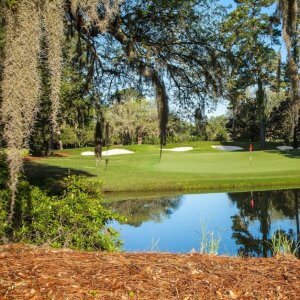 A picturesque view of Caledonia Golf & Fish Club in Pawleys Island, featuring a green framed by moss-draped oaks, sand bunkers, and a reflective water hazard.