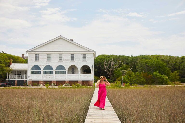 A woman in a pink dress walks along a wooden boardwalk through a marsh with a white house in the background