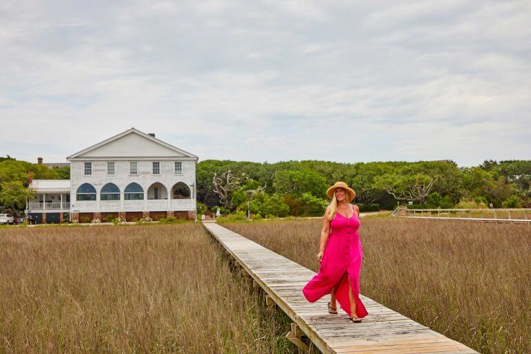 A woman in a pink dress walks along a wooden boardwalk through a marsh with a white house in the background