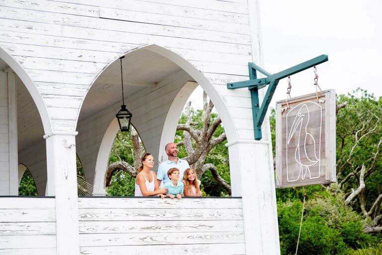 A family looks out from a white arched balcony next to a wooden pelican sign