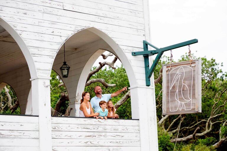 A family looks out from a white arched balcony next to a wooden pelican sign