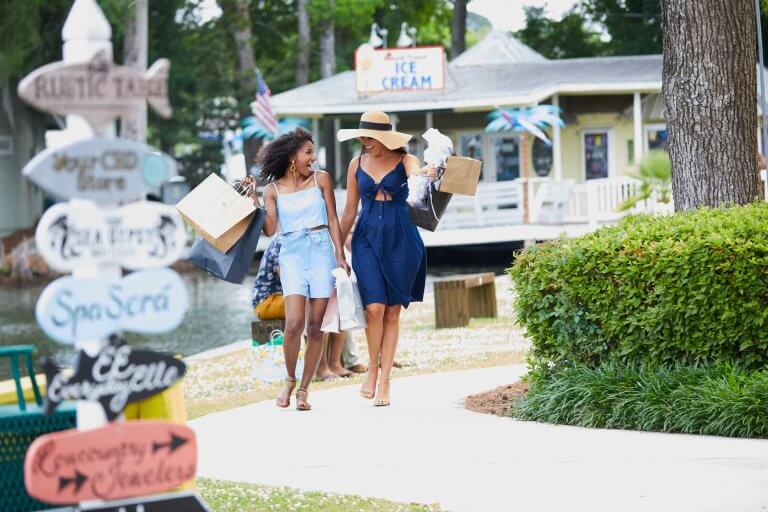 Two women laugh while carrying shopping bags past a row of colorful shops on Pawleys Island, SC
