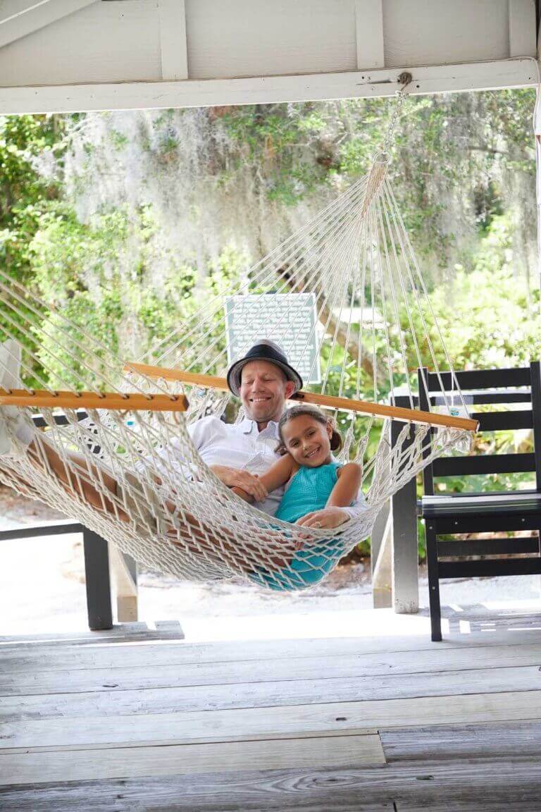 A man and a young girl smile while relaxing together in a rope hammock on a porch