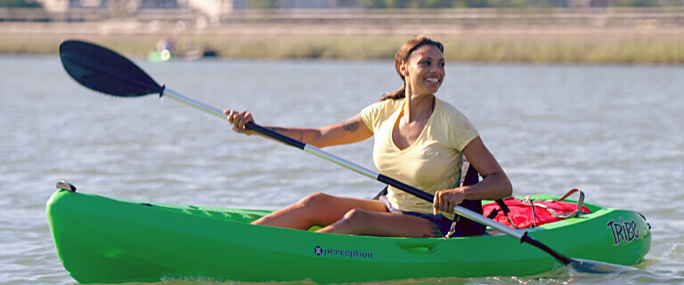A smiling woman paddles a bright green kayak on calm water around Pawleys Island, SC