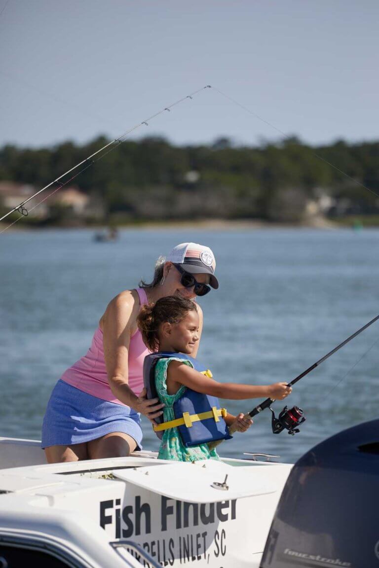 A woman helps a young girl in a life jacket fish from a boat on a sunny day
