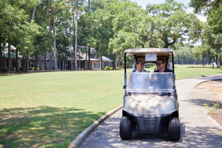 A man and woman laugh while driving a golf cart on a sun-dappled Pawleys Island golf course