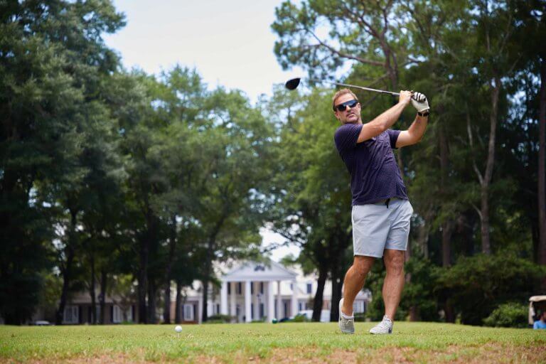 A man in sunglasses completes a golf swing on a green with a white clubhouse in the background