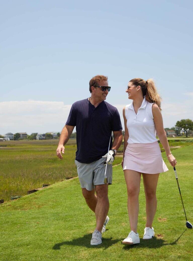 A man and a woman smile at each other while walking with golf clubs on a green course next to a marsh