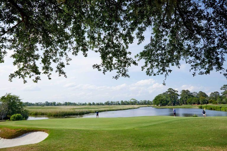 Golfers on a green overlooking a scenic marsh and waterway, framed by tree branches