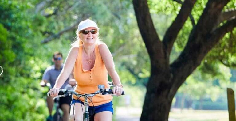 A woman in a sun hat and sunglasses smiles while riding a bicycle on a tree-lined path